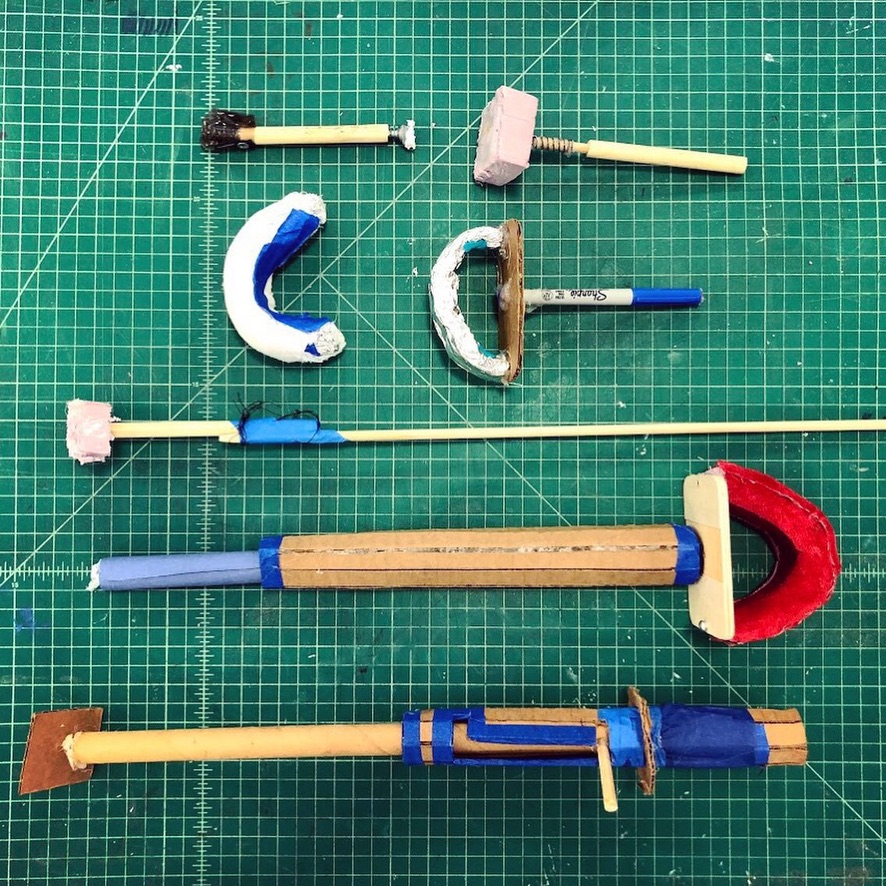 Collection of prototypes for a button-pressing assistive device on a green gridded background. The first two devices are short sticks with foam-like tips. The second two below them have c-shaped curved handles. The next one is a long stick with foam on the end. The next is scored cardboard with a velvet and foam handle. The last is scored cardboard with a groove in the side that allows extension of the length of the device and also has a swiveling flat tip.