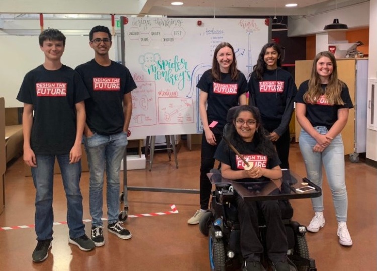 Photograph of Sofia and 5 students in black t-shirts with red and white word reading "Design the Future". In the foreground, one student in a motorized wheelchair holds up the design prototype, an extendable button pusher, smiling. Just behind her, the other students pose and smile. In the background, there is a whiteboard with the design team name, "Spider Monkeys", and some partially obscured drawings of different prototypes and stages in design thinking.
