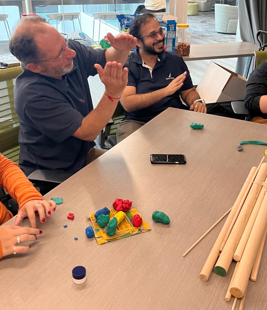 Two co-design day participants smile as one expresses an idea using gesture. Another participant's hands reach for colorful clay on the table.