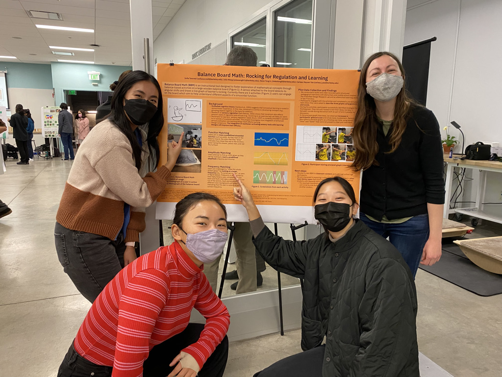 The balance board math design team poses cheerfully surrounding a bright orange poster.