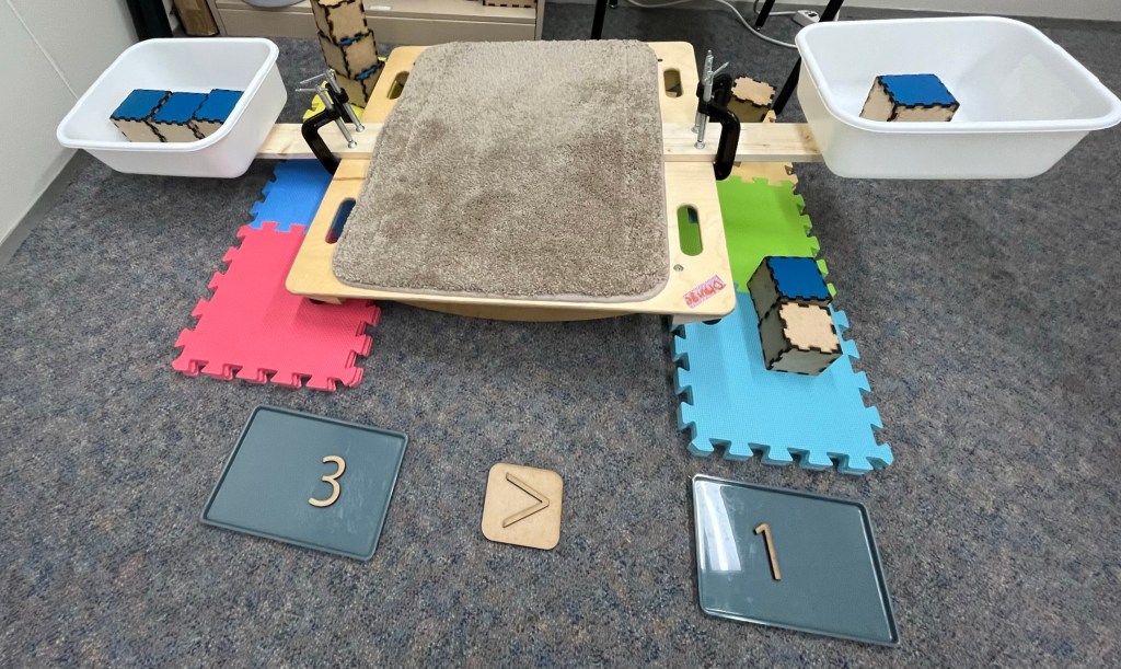 Photograph of a balance board with wooden beams extending from each side. At the ends of the beams are plastic bins containing weighted wooden blocks. In front of the board, wooden numerals document the number and comparison of the amounts on either side of the bin: 3 > 1. 