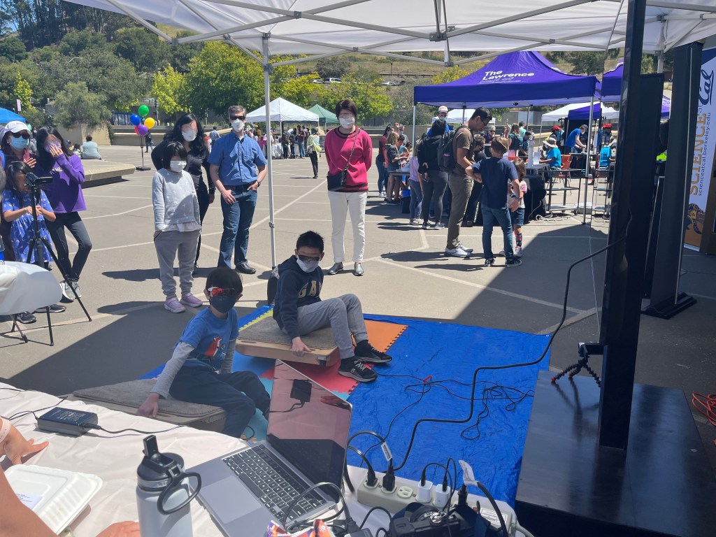Two children in masks who look to be elementary age rock on balance boards, with a crowd of observers and visitors milling behind them.