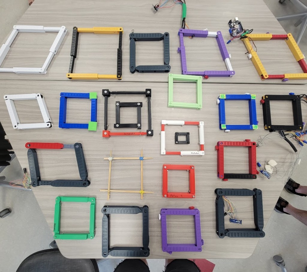 Photograph of about 20 different quadrilateral prototypes of assorted sizes and styles, ranging from a set of chopsticks rubber-banded together to 3d-printed, wired sensing-acutuating versions.