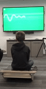 a child rocks front to back on a wooden balance board, generating a digital graphs on a screen in front of him.