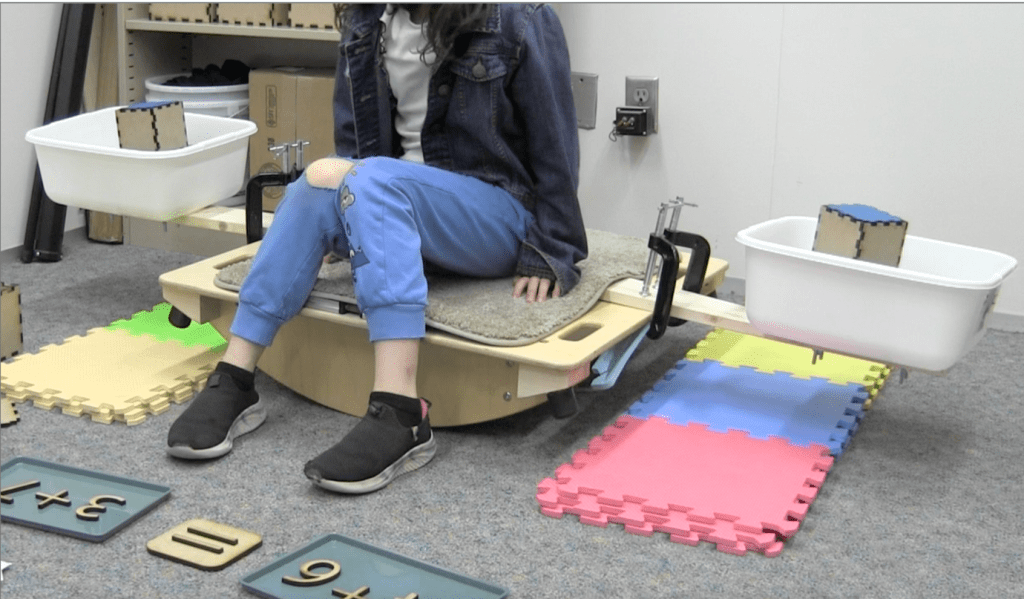 Photograph of a child sitting on a wooden balance board with wooden beams extending from each side. At the ends of the beams are plastic bins containing weighted wooden blocks. In front of the board, wooden numerals document the amounts being compared on either side of the bin 4+6 = 3+7.