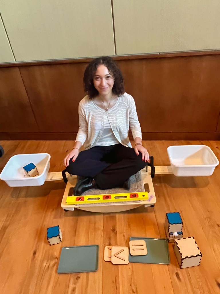 BBM team member Amanda Alegria sits on the Equations and Inequalities balance board with bins extending from each side. She has a single block on one side, which is tilting the board and her body to that side.