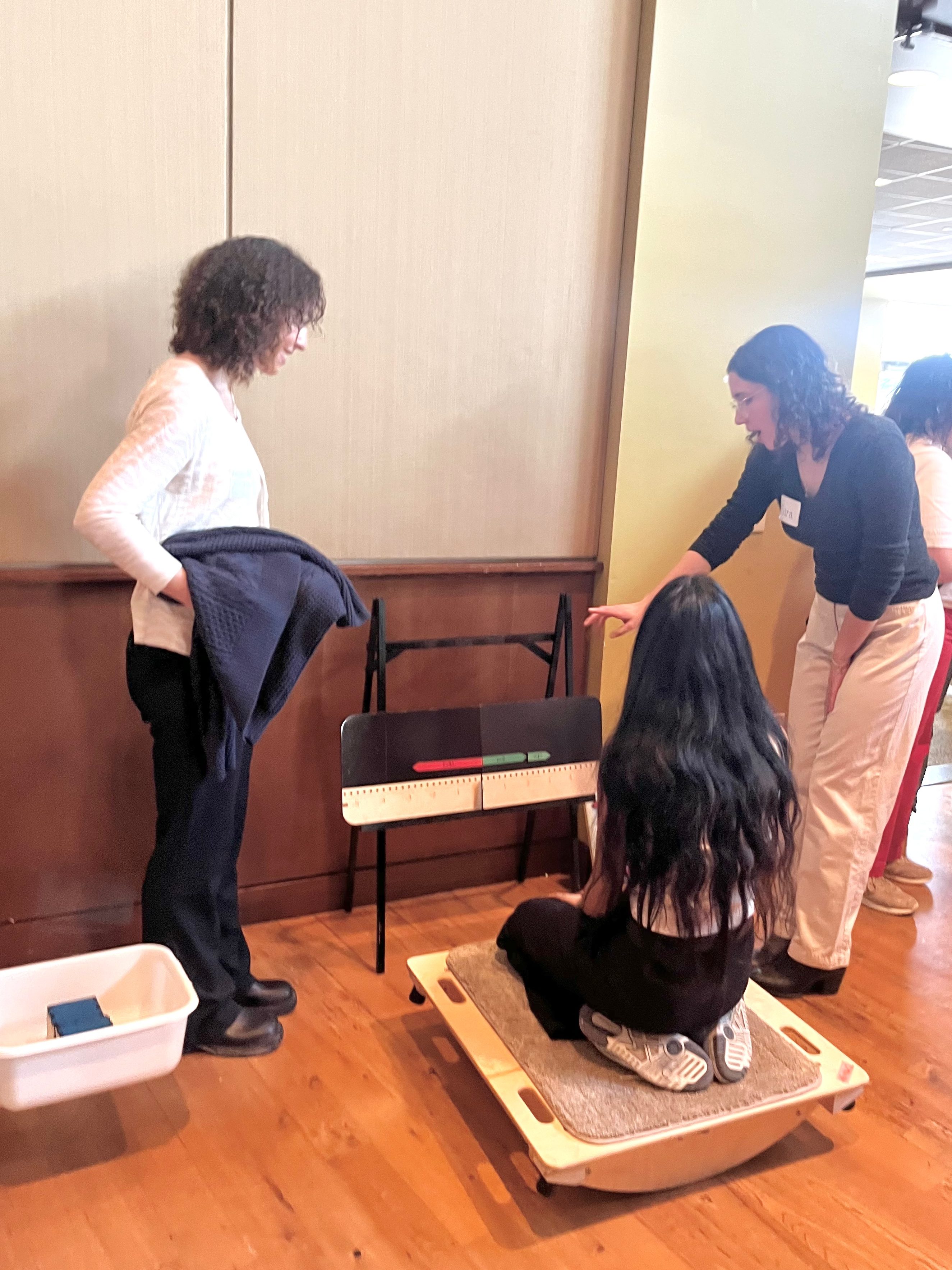 A demo visitor kneels on a balance board in front of a number line on an easel while 2 project team members invite them to try different tasks.