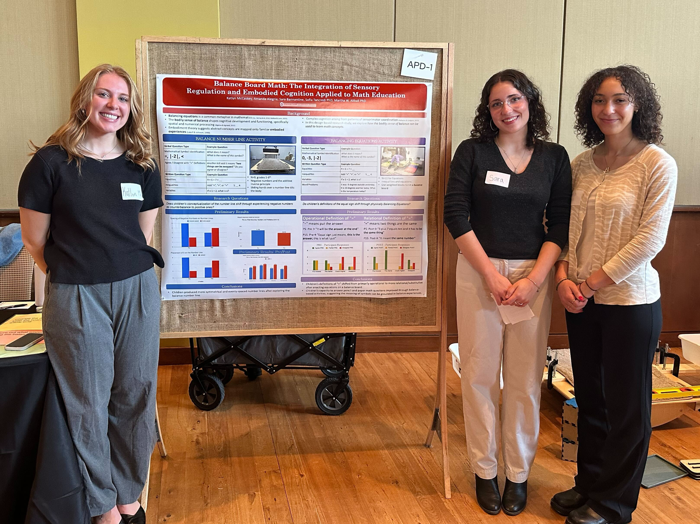 Three undergraduate researchers smile in front of a research poster with a balance board activity demo in the background.