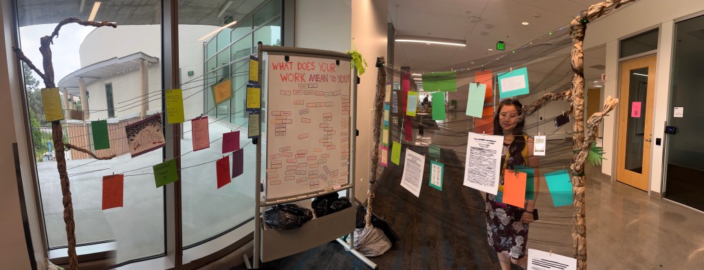 An installation of researchers' poems clipped onto strings strung among different wood and paper trees. In the middle, there is a whiteboard of magnetic poetry with the question, 